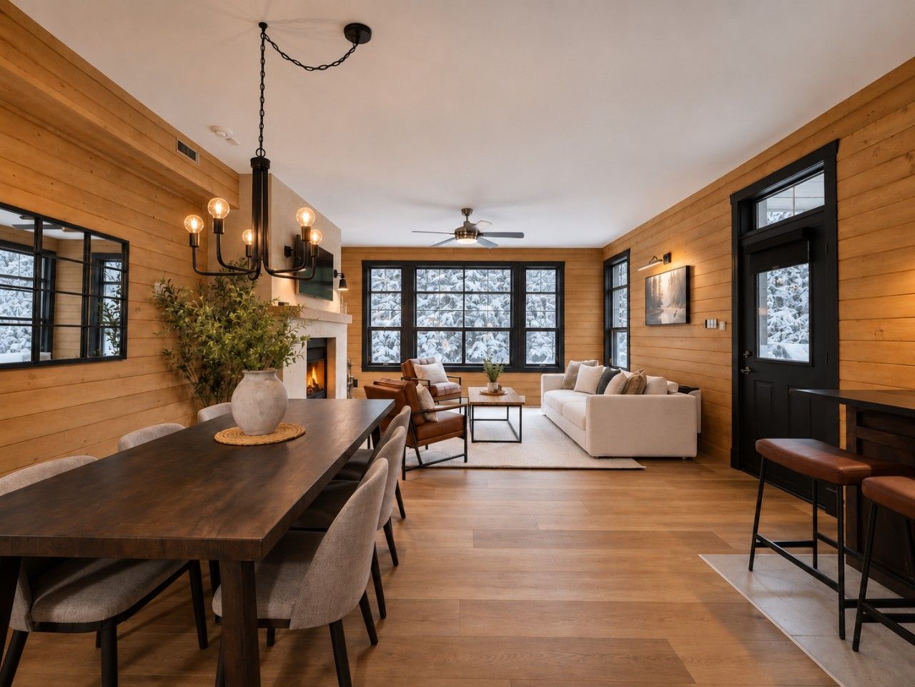 A modern interior with a wooden floor, a dining area with a table and chairs, a sofa set, and large windows with a view of snow-covered trees.