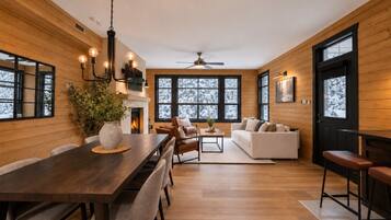 A modern interior with a wooden floor, a dining area with a table and chairs, a sofa set, and large windows with a view of snow-covered trees.
