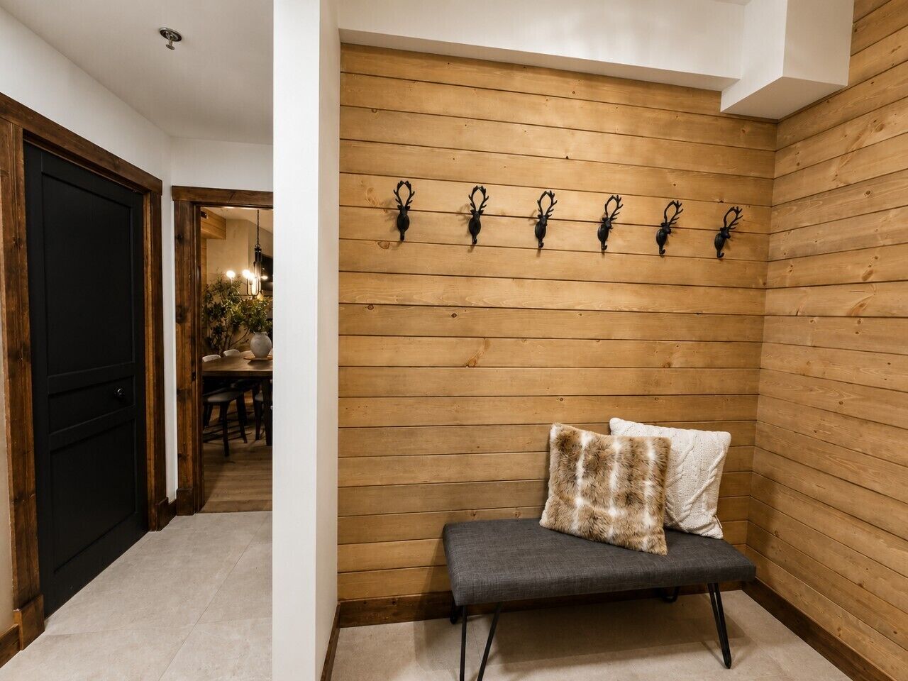 Un mur lambrissé en bois, avec un banc et des coussins, une porte noire et, au fond, un coin repas.