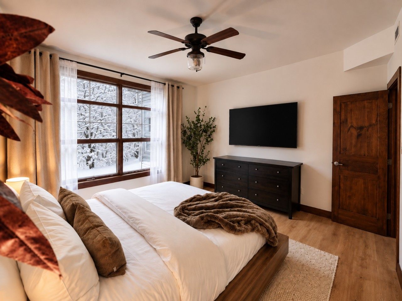 Une chambre à coucher comprenant un lit, un ventilateur de plafond, un téléviseur fixé au mur, une commode en bois et une porte.