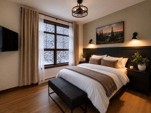 Ein Schlafzimmer mit einem Bett, einer Bank, einem Nachttisch, einem an der Wand montierten Fernseher, einem Gemälde und einem Fenster mit Vorhängen.