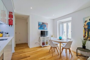 Eine moderne Küche mit weißen Schränken, ein Essbereich mit einem runden Tisch und Stühlen, ein Fernseher auf einem weißen Schrank und ein Bild an der Wand.