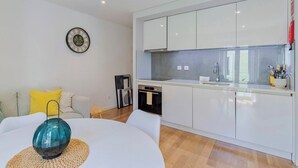 A modern kitchen with white cabinets, a built-in oven, and a sink. There is a dining table with a glass vase and a woven placemat. A sofa with yellow cushions is in the living area. - Blue Coconut Second, By Home Match (Setúbal)