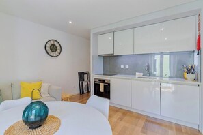 Eine moderne Küche mit weißen Schränken, einem eingebauten Ofen und einem Spülbecken. Ein Esstisch mit einer Glasvase und einer gewebten Tischmatte. Im Wohnbereich steht ein Sofa mit gelben Kissen.