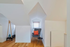 A loft-style room with a sloped ceiling, a black and white striped chair with an orange cushion, a small black stool, and a wooden swing. - Blue Coconut Second, By Home Match (Setúbal)