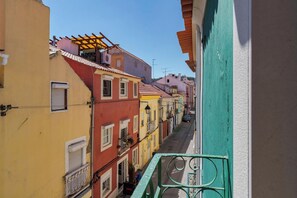 A narrow street with colorful buildings, a balcony with a metal railing, and a clear blue sky. - Blue Coconut Second, By Home Match (Setúbal)