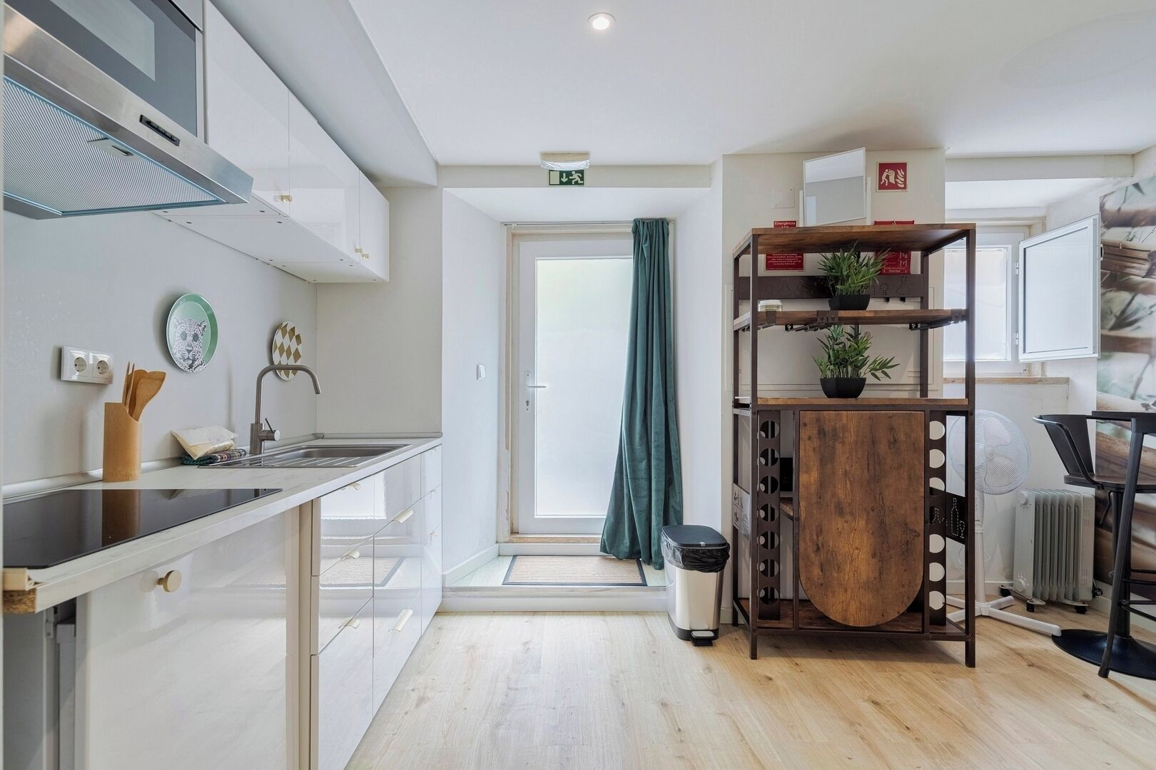 A modern kitchen with a stainless steel range hood, a sink, and a countertop with kitchen utensils. There is a wooden shelf with a plant and a small table with a chair.