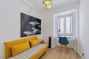Ein Zimmer mit einem gelben Sofa, einem Schreibtisch mit Stuhl, einem großen Schwarz-Weiß-Kunstwerk und einem Fenster mit Jalousien.