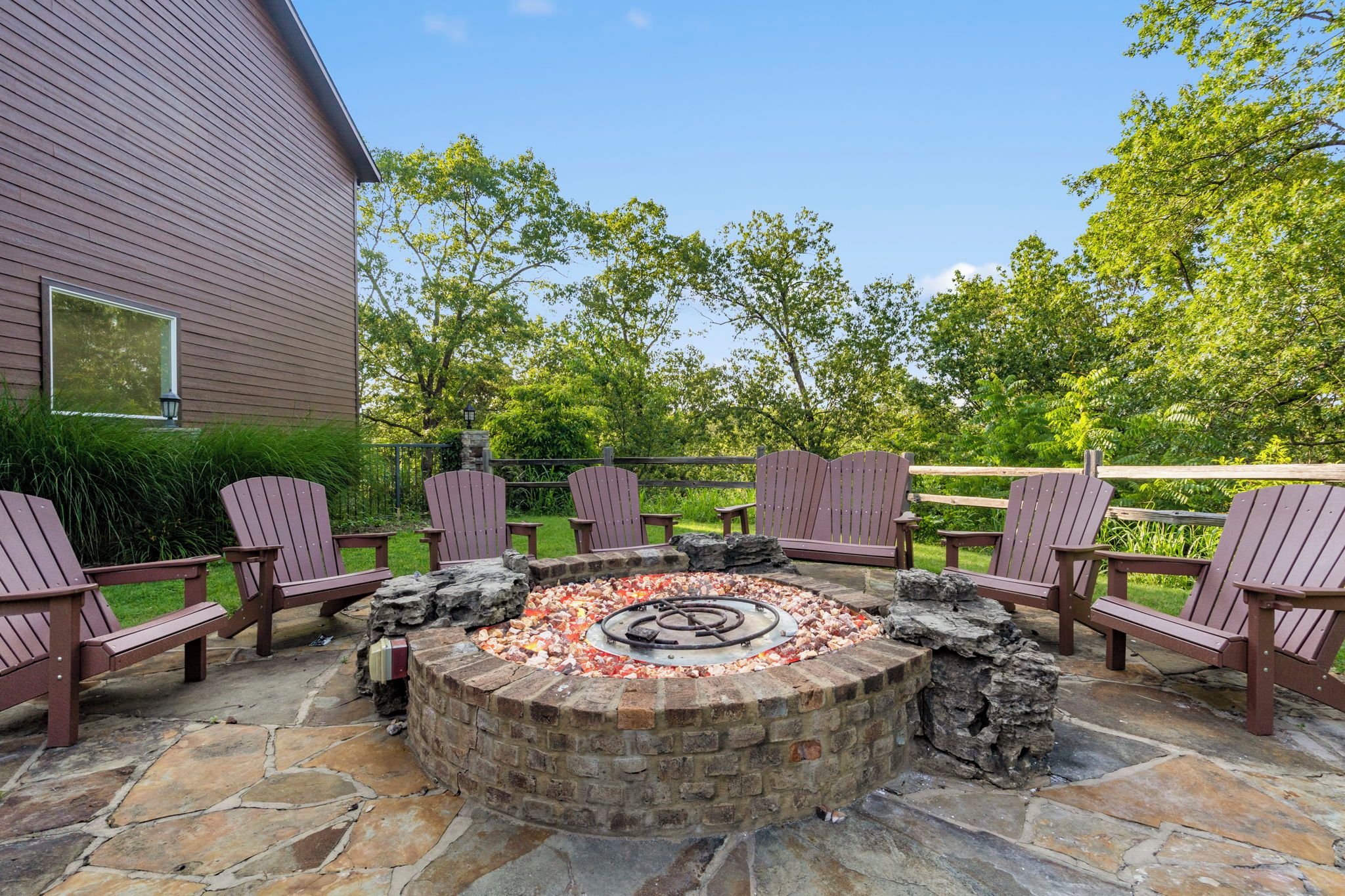 A circular stone fire pit surrounded by Adirondack chairs, set on a stone patio with a wooden fence and greenery in the background.