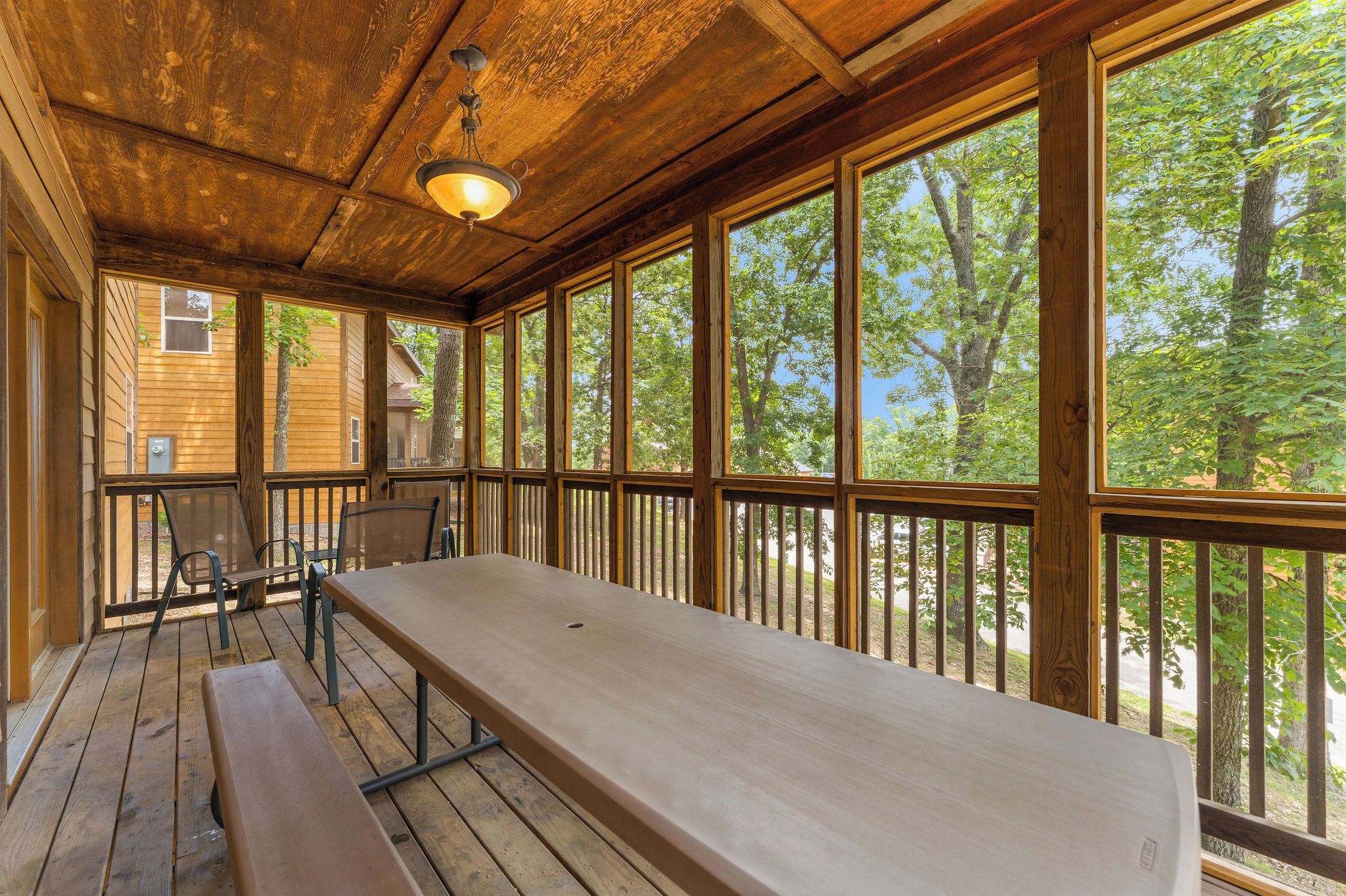 A wooden deck with a long table and benches, large windows with a view of trees, and a hanging light fixture.
