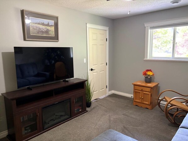 A living room with a flat-screen TV on a wooden stand, a door, a window with a view, and a small wooden cabinet with a vase of flowers.
