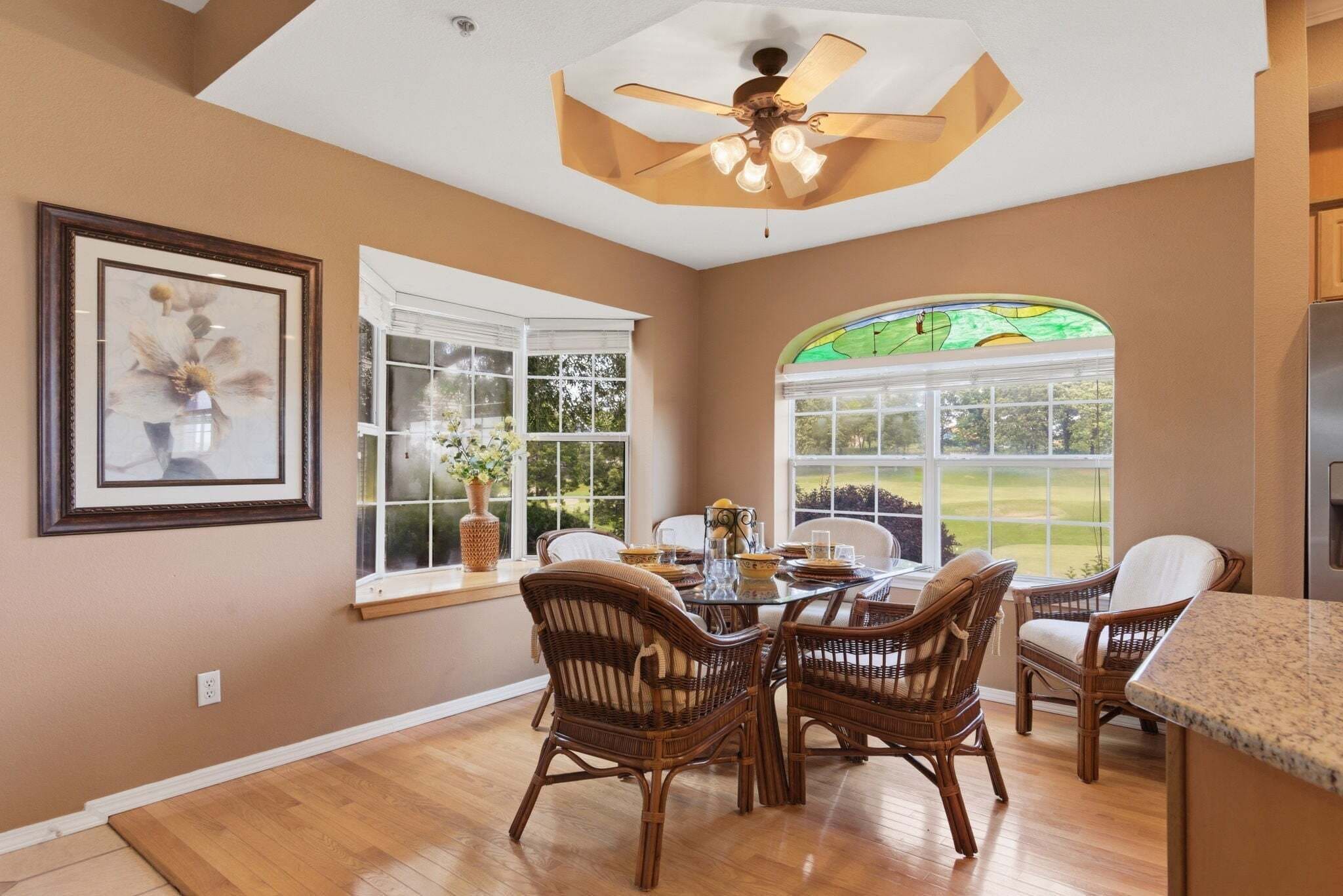 A dining area with wicker chairs, a ceiling fan, and large windows with a view of a green landscape.