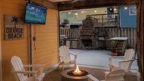 Un espacio de estar al aire libre con techo, sillones Adirondack, un brasero, un televisor montado en la pared y una chimenea de piedra.
