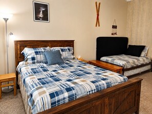 Ein Schlafzimmer mit einem hölzernen Bett, einem zusätzlichen Bett, einem Nachttisch, einer Lampe, einem gerahmten Bild und einer Wanddekoration.
