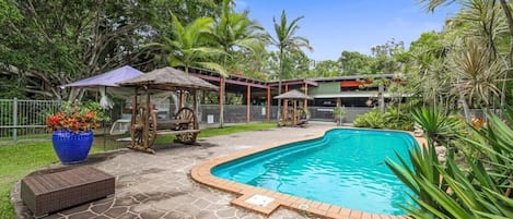 Una piscina con un patio de piedra, rodeada de vegetación tropical y un pabellón.
