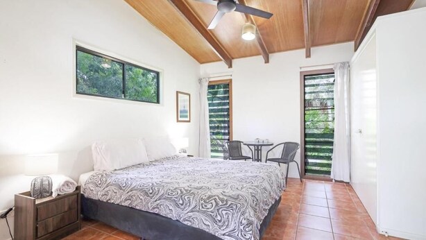 Un dormitorio con cama, ventilador de techo, ventana, puerta y una pequeña mesa con sillas.
