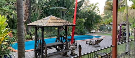 Una zona de piscina con una estructura de madera, sillas de descanso y un jardín cercado.