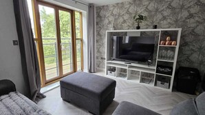 A modern living room with a TV, grey sofa, ottoman, and a balcony with a view.