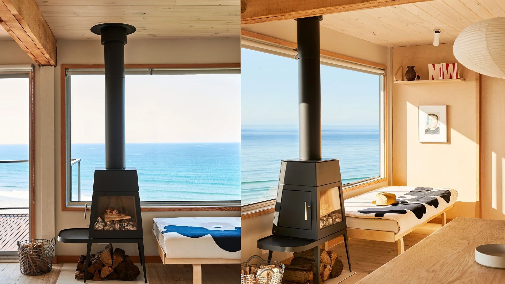 A room with a large window, a black wood-burning stove, a bed with a blue blanket, and a wooden table.