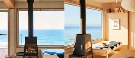 A room with a large window, a black wood-burning stove, a bed with a blue blanket, and a wooden table.