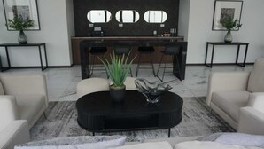 Uma sala de estar moderna com uma mesa de centro preta, duas plantas em vasos, um sofá e uma área de cozinha com balcão.