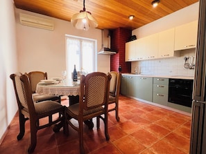 A dining area with a table set for four, wooden chairs, a chandelier, and a kitchen with white cabinets and a red brick wall. - A6 - Spacious apt w pool, terrace and sea view (Dugi Rat)