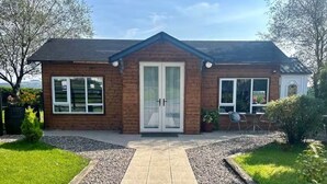 A two-story wooden house with a front door, two windows on the ground floor, and a small patio area with a table and chairs. - Gortin Glen Forest Cabin, Sleeps 7 with Parking (Omagh)