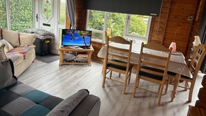 A wooden cabin interior with a TV on a stand, a sofa, a dining table with chairs, and a view of greenery outside. - Gortin Glen Forest Cabin, Sleeps 7 with Parking (Omagh)