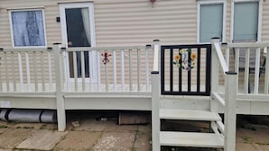 En mobilhome med hvide vægge, en veranda med hvidt gelænder, og et lille haveområde med en velkommenskilt.