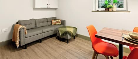 A living room with a grey sofa, a small table with orange chairs, a wooden floor, and a window with plants.