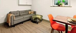 A living room with a grey sofa, a small table with orange chairs, a wooden floor, and a window with plants. - GuestReady - Studio w Kitchenette in Harrow (London)