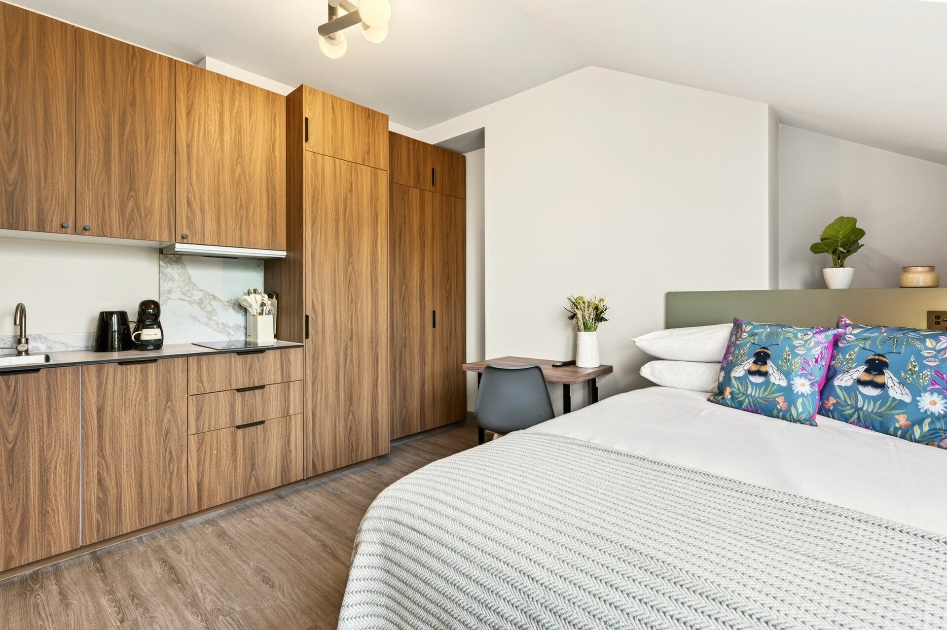 A modern bedroom with a large bed, a wooden wardrobe, a small table with a vase, and a nightstand.