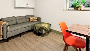 A living room with a grey sofa, a small table with orange chairs, a wooden floor, and a window with plants. - GuestReady - Trendy Studio in Harrow (London)
