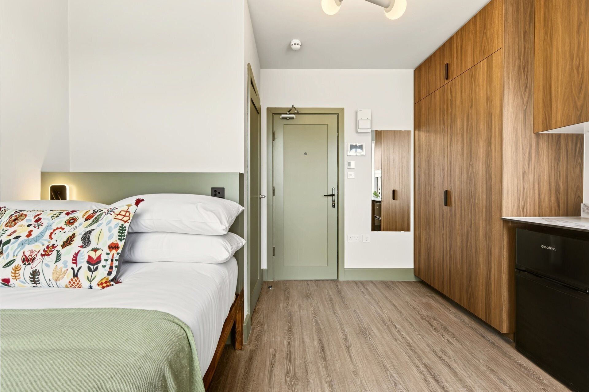 A bedroom with a bed, a bedside table, a wardrobe, and a kitchenette area.