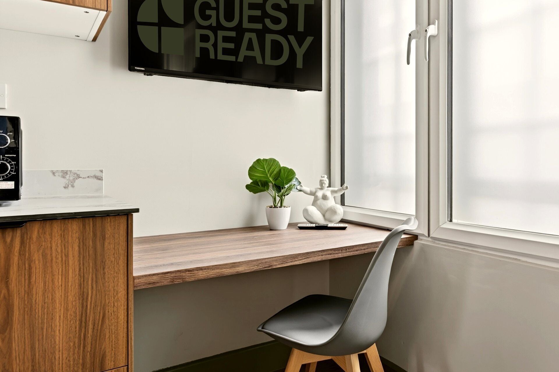 A modern room with a wooden desk, a chair, a potted plant, a decorative sculpture, and a wall-mounted TV displaying 'GUEST READY'.