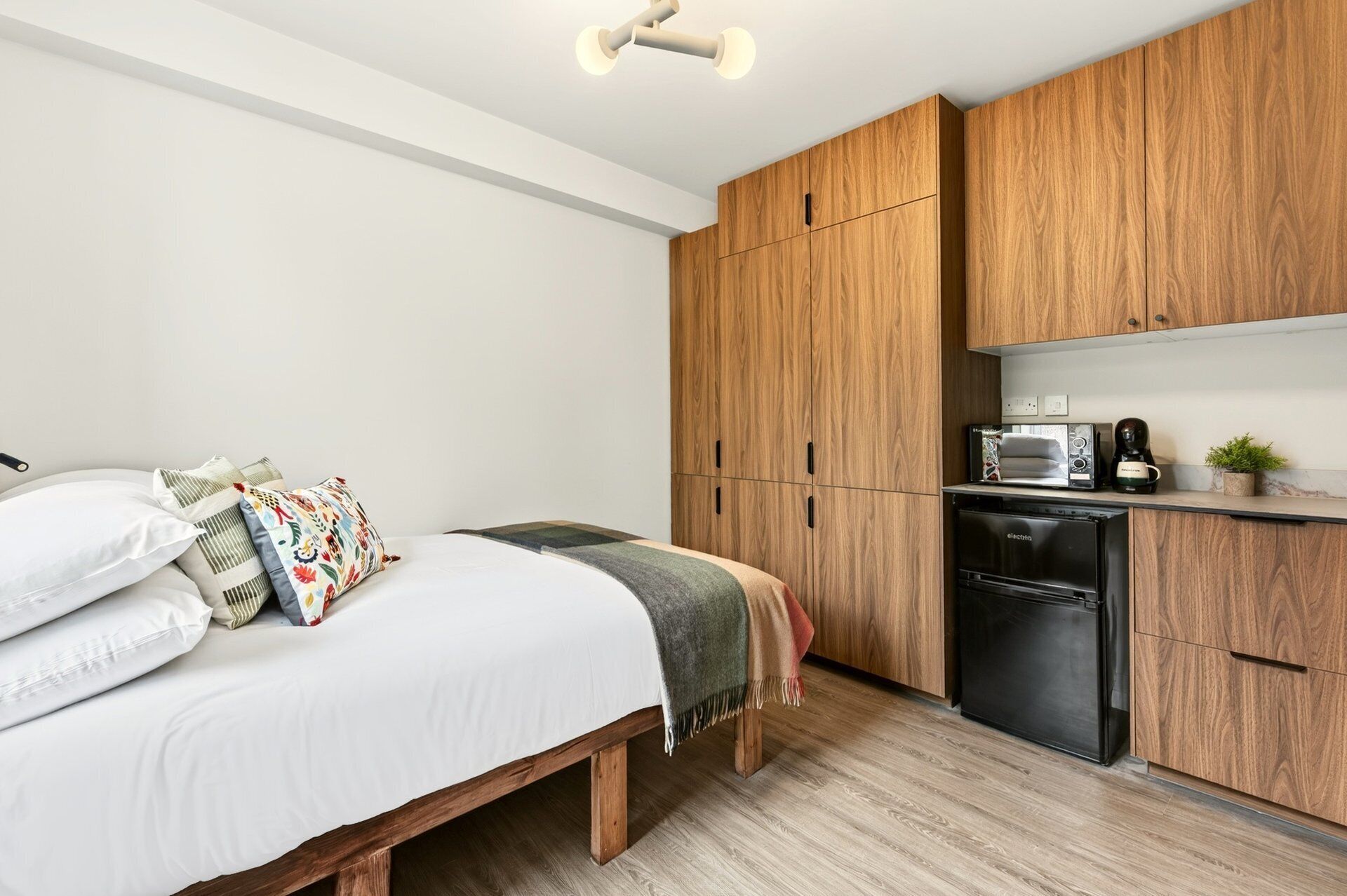A compact bedroom with a bed, bedside table, and a small kitchenette with a microwave and refrigerator.