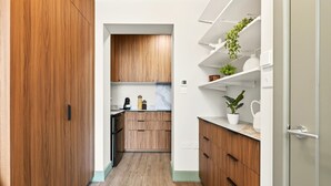 Ein schmaler Flur führt zu einer Küche mit Holzschränken, einer Marmorarbeitsplatte und weißen Regalen mit Pflanzen.