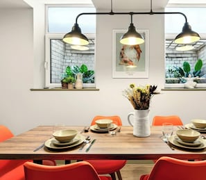 Comedor con mesa de madera para cuatro personas, sillas naranjas y lámparas colgantes.