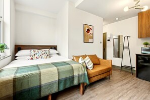 Quarto moderno com cama, sofá, pequena cozinha e banheiro visível por uma porta aberta.