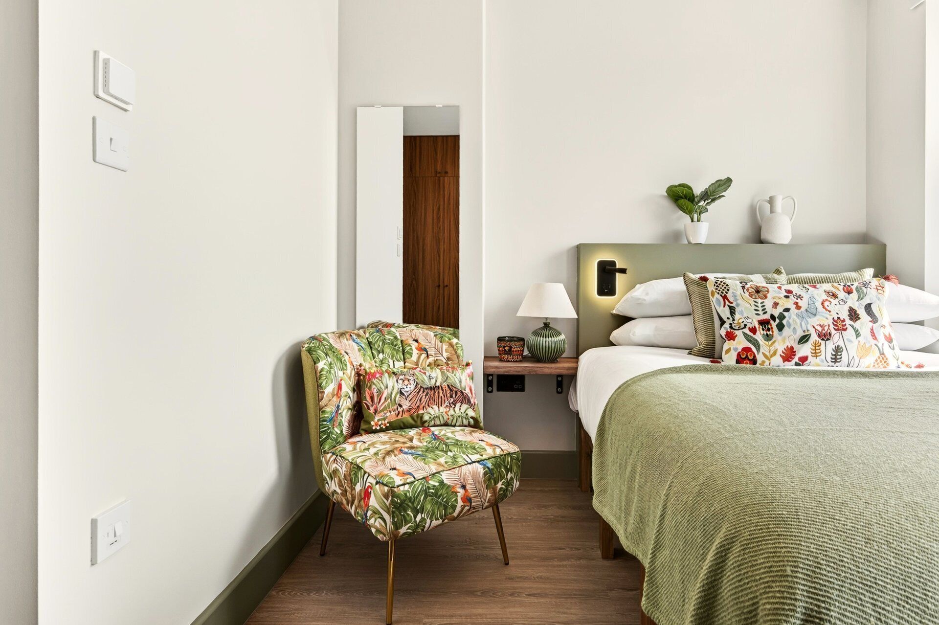 Une chambre d’hôtel avec un lit, une chaise, une table de chevet, une lampe et une plante.