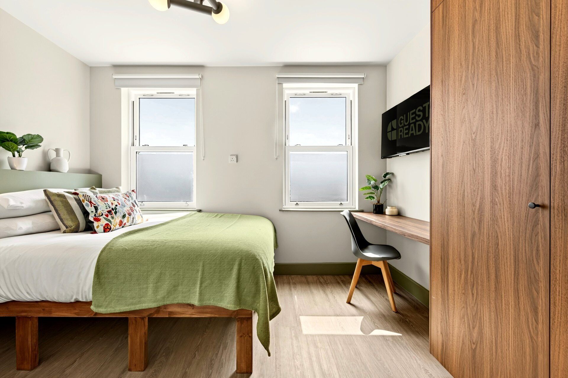 A bedroom with a bed, two windows, a desk, a chair, a plant, and a wooden wardrobe.