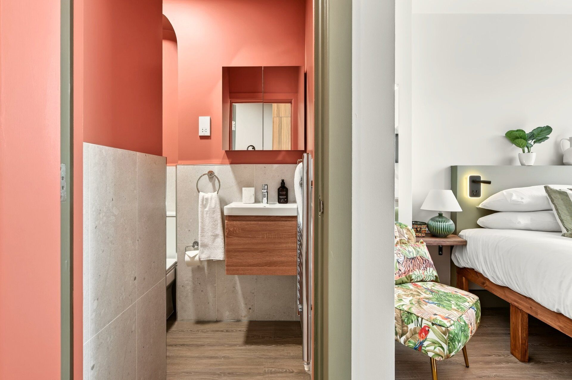 A hotel room with a wooden floor, a bed with white bedding, a nightstand with a lamp, a small table with a plant, and a bathroom with a wooden vanity and a towel rack.