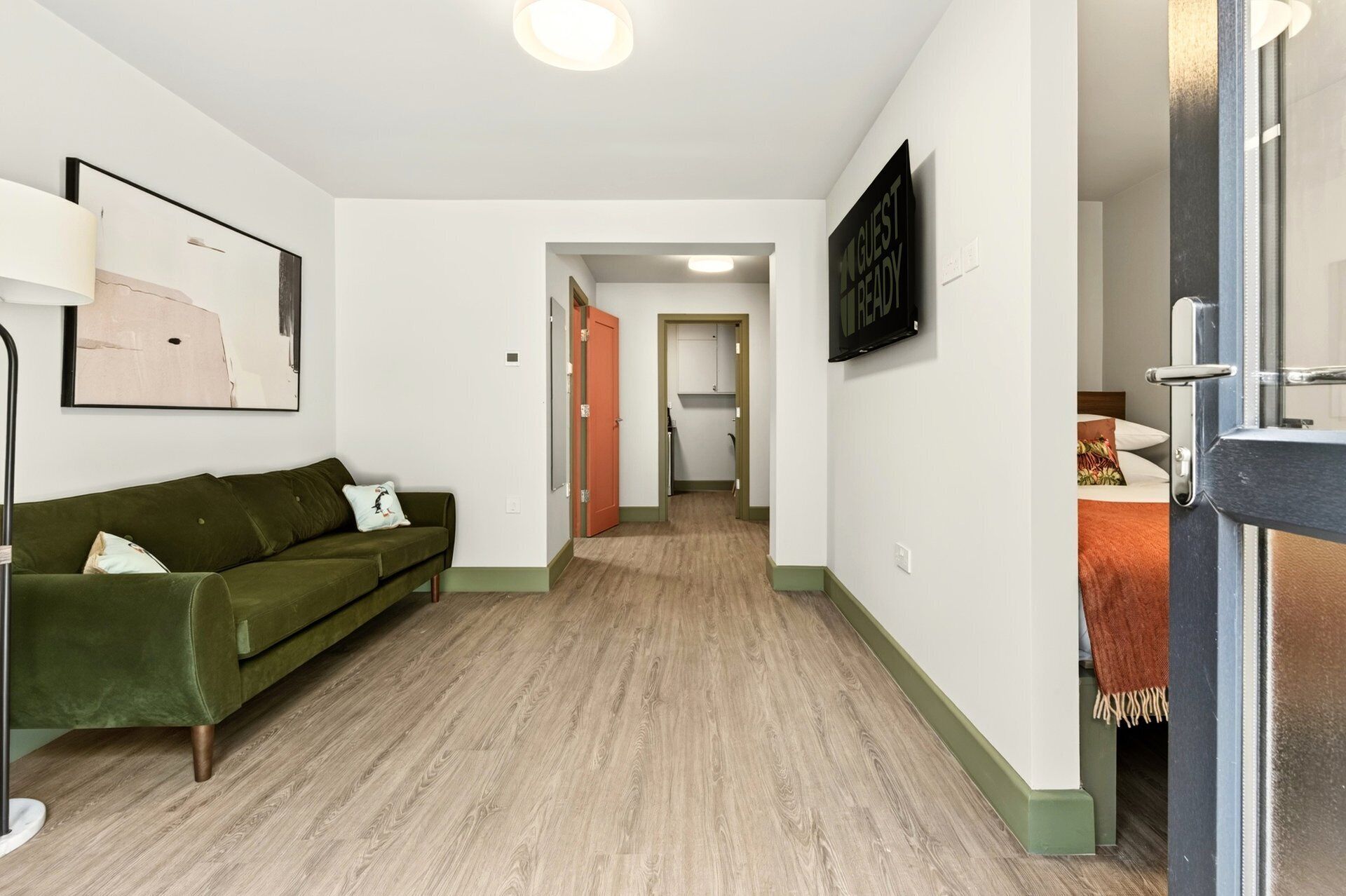 Ein Hotelkorridor mit einem grünen Sofa, einem gerahmten Kunstwerk, einem Flachbildfernseher und Holzboden.