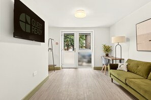 A modern living room with a green sofa, a TV mounted on the wall, and a sliding glass door leading to a balcony. - GuestReady - Modern 1BR w Full Kitchen in Harrow (London)