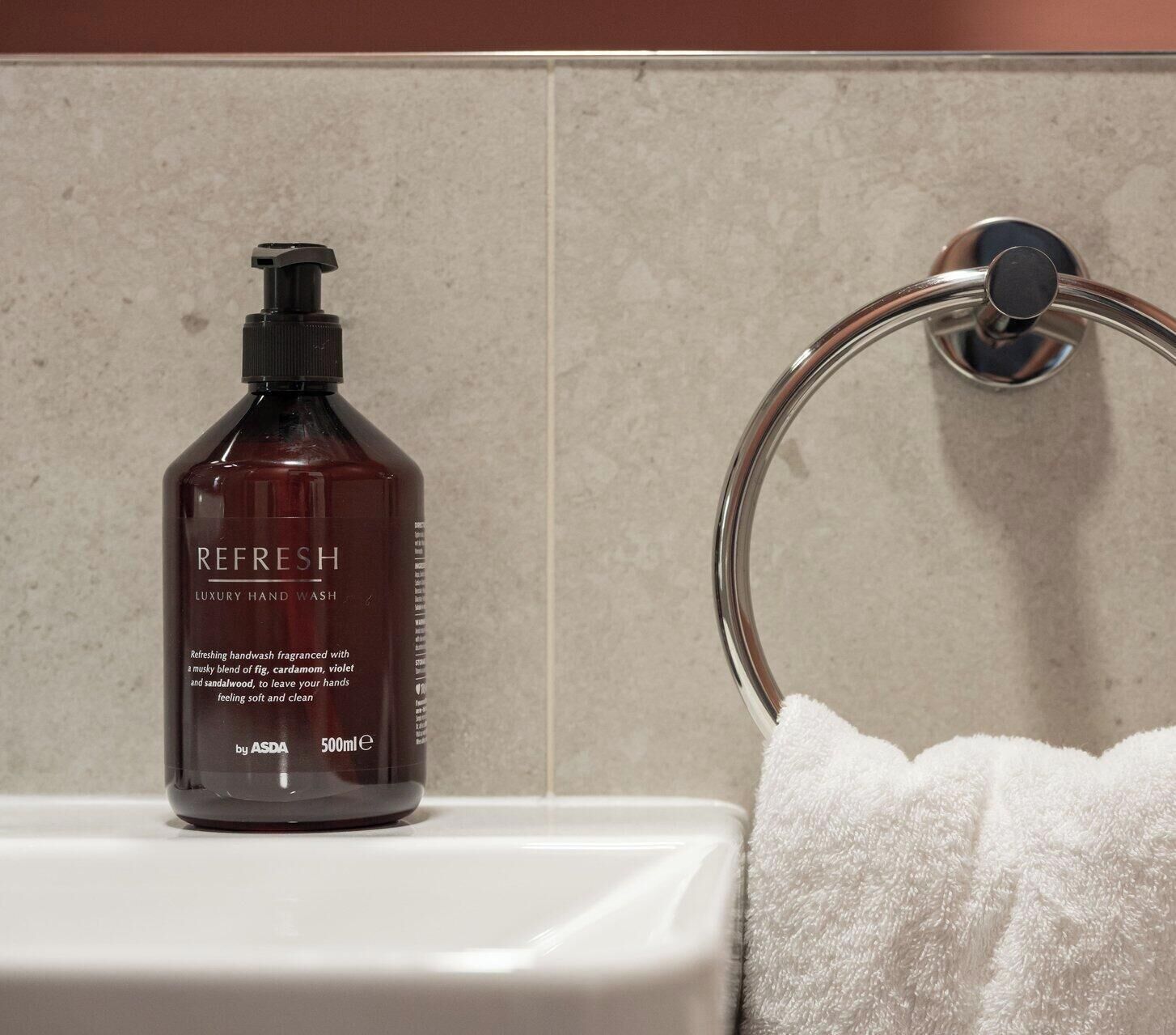 Une salle de bain équipée d’un distributeur de savon liquide portant l’étiquette « SAVON POUR LES MAINS DE LUXE – RAFRAÎCHISSANT » de la marque ASDA, d’un porte-serviette et d’une serviette blanche.