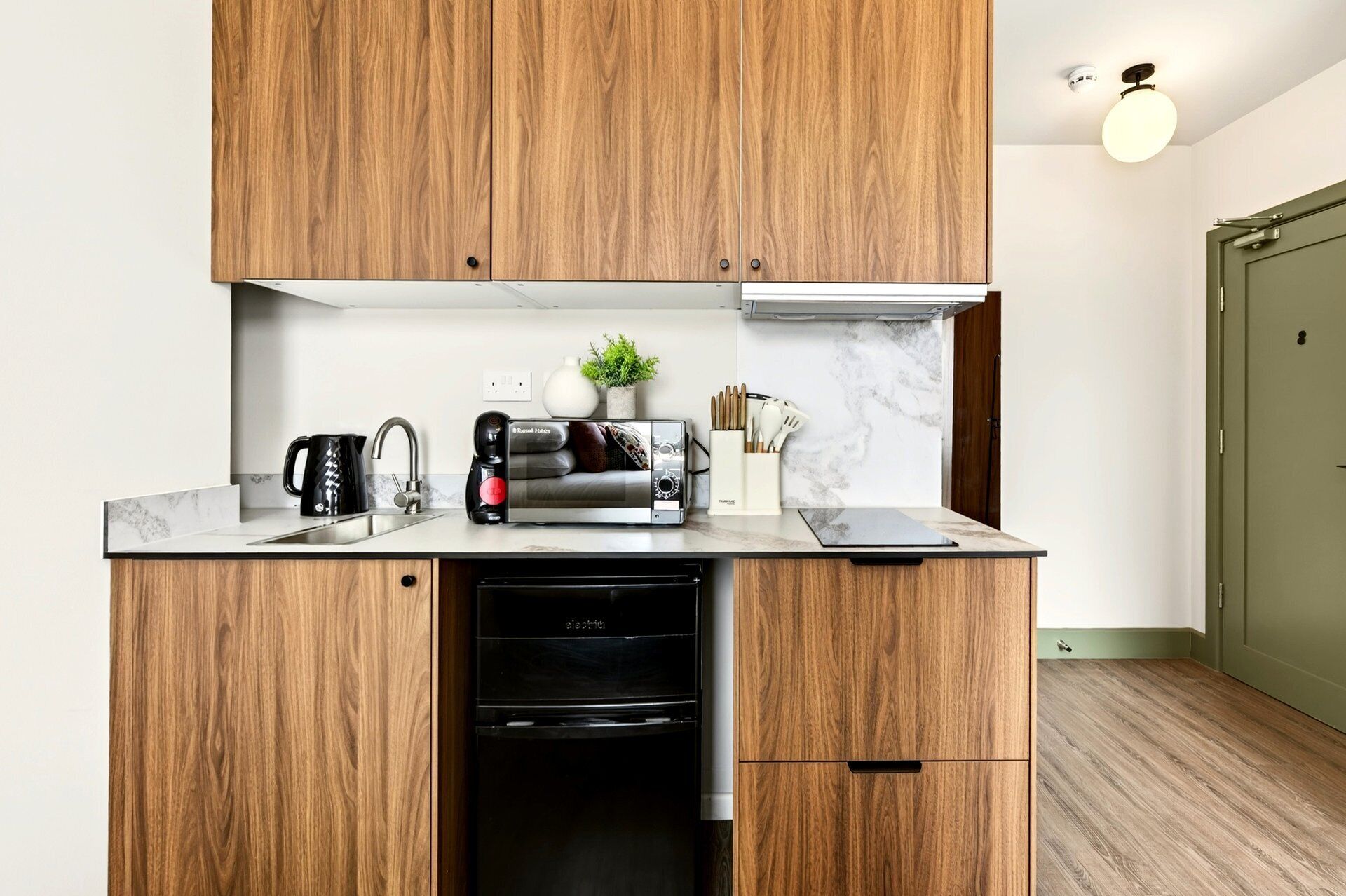 Cocina compacta con armarios de madera, lavavajillas integrado, cafetera y una pequeña planta en maceta.