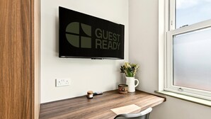 A modern room with a wooden desk, a mounted TV displaying 'GUEST READY', a small potted plant, and a window with a view. - GuestReady - Chic Studio w Kitchenette in Harrow (London)
