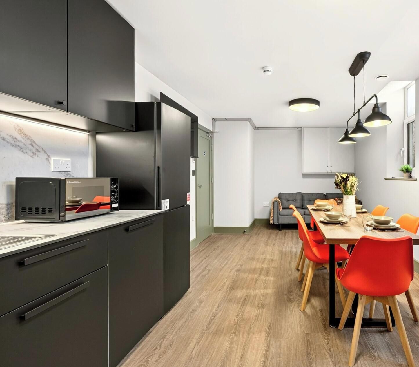 A modern kitchen with a built-in microwave, a dining area with a wooden table and red chairs, and a grey sofa in the background.