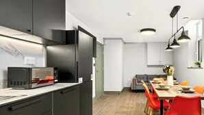 Uma cozinha moderna com micro-ondas embutido, uma sala de jantar com mesa de madeira e cadeiras vermelhas, e um sofá cinza ao fundo.