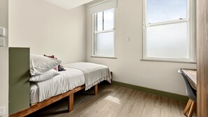 Quarto pequeno com uma cama de solteiro, chão de madeira, duas janelas e uma luminária no teto.
